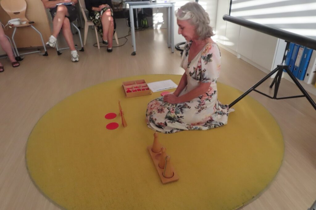 Renée Classen zeigt auf dem Teppich Montessori-Material, Workshop-Teilnehmende sitzen im Halbkreis